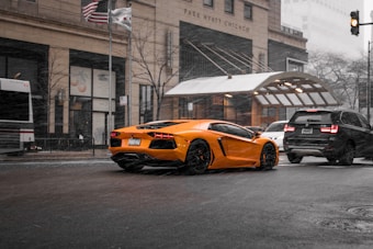 A vibrant orange sports car is driving through a city street amidst a snowstorm. Surrounding vehicles include a bus and a black SUV. The background features the Park Hyatt Chicago building, adorned with American and Chicago flags.