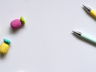 Two colorful pens are positioned diagonally in the top right corner on a plain white background. To the left, there are two erasers shaped like pineapples, one pink and the other yellow, each with a green leafy top.