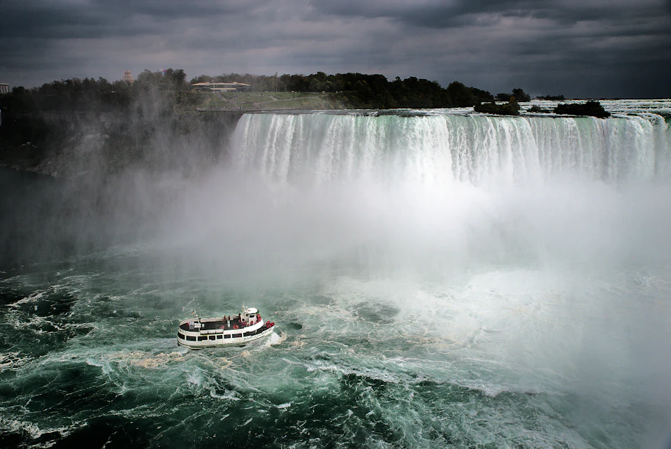 The image depicts a white boat with red accents, likely a tour boat or ferry, sailing on the water near a waterfall. The boat is positioned towards the right side of the frame and appears to be moving away from the viewer. In the background, there are trees visible in the distance, adding depth to the scene. The sky above is filled with clouds, suggesting an overcast day.