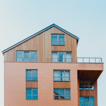 A modern building with a geometric design featuring a combination of brick and wood paneling. The structure has multiple large windows that reflect the sky, and there is a balcony area with a glass railing.