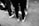 A group of people stand on a rough surface with a focus on their footwear. They are wearing various styles of sneakers, and one person has a drink can attached to their bag. The image is in black and white.