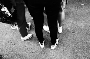 A group of people stand on a rough surface with a focus on their footwear. They are wearing various styles of sneakers, and one person has a drink can attached to their bag. The image is in black and white.