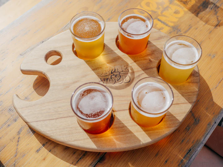 Cinco copos de cerveja de cores e estilos diferentes lado a lado em tábua de degustação, luzes douradas ao fundo