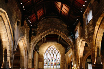 The interior of an old English church with stone arches, wooden pews and a stained glass window