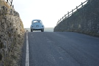 A classic car is driving along an empty road nestled between two stone walls. The scene is captured from behind, showcasing the car's rear lights and license plate. The road has a gentle uphill gradient, and the walls are topped with wooden fences. The landscape is rural, with dry grass lining the stone walls.