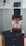 A vape device featuring a black and silver cylindrical tank mounted on a sleek black base. The tank displays glass and metal components with visible branding. The background is softly blurred, highlighting a modern, minimalistic setting.