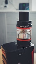 A vape device featuring a black and silver cylindrical tank mounted on a sleek black base. The tank displays glass and metal components with visible branding. The background is softly blurred, highlighting a modern, minimalistic setting.