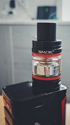 A vape device featuring a black and silver cylindrical tank mounted on a sleek black base. The tank displays glass and metal components with visible branding. The background is softly blurred, highlighting a modern, minimalistic setting.