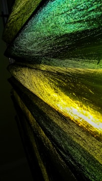 The image features several layers of translucent, textured fabric in shades of green and yellow. The light filters through the material, creating a glowing effect that highlights the intricate patterns and textures within the fabric. The colors transition smoothly from deep green to bright yellow, adding depth and movement.
