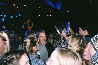 A crowd of people, mostly young adults, are gathered at what appears to be a lively event, possibly a party or concert. One person is holding a large transparent container, and some people are talking and engaging with each other. The scene has dim lighting with spotlights and people appear energetic and animated.