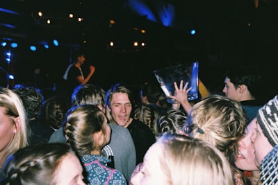 A crowd of people, mostly young adults, are gathered at what appears to be a lively event, possibly a party or concert. One person is holding a large transparent container, and some people are talking and engaging with each other. The scene has dim lighting with spotlights and people appear energetic and animated.