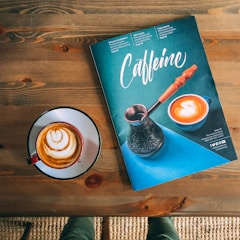 A magazine titled 'Caffeine' with an image of a coffee cup and a cezve is placed on a wooden table. Next to it, there is a cup of coffee with latte art.