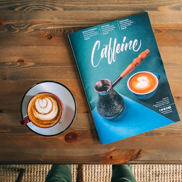 A magazine titled 'Caffeine' with an image of a coffee cup and a cezve is placed on a wooden table. Next to it, there is a cup of coffee with latte art.