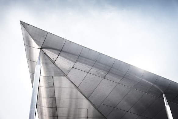 A modern architectural structure featuring a large, angular roof made of metal panels. The design emphasizes clean lines and geometry, and the bright sky offers a contrasting backdrop, accentuating the metallic sheen.