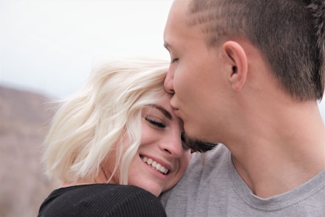 A couple shares an intimate moment where a man gently kisses the forehead of a smiling woman with short blonde hair. She leans into him, both displaying a sense of affection and happiness. They are dressed casually, and the background is an out-of-focus, natural setting.