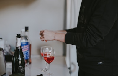 A person is preparing drinks, pouring a red beverage into wine glasses. Bottles, including what appears to be wine and spirits, are present on a countertop. The background is softly focused, emphasizing the activity.
