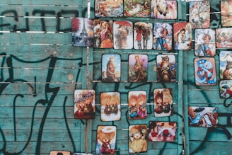 A collection of colorful, whimsical artworks mounted on a weathered, turquoise wooden fence with black graffiti. The artworks include expressive illustrations of animals, fantasy creatures, and people in various styles and settings. Each piece is unique, showcasing vibrant colors and imaginative scenes.
