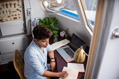 A person is sitting at a wooden desk in a cozy room, writing on a notepad while looking at a laptop. There are various items on the desk, including a closed notebook, a potted plant, a glass jar, a lamp, and an alarm clock. A window provides natural light, and there is a decorative board with illustrations of crustaceans on the wall.