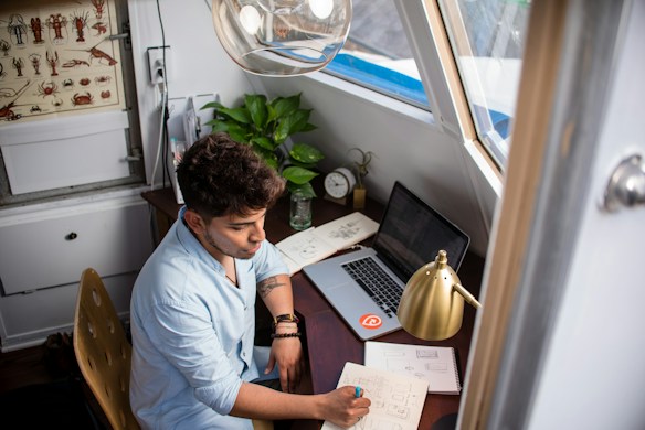 A person is sitting at a wooden desk in a cozy room, writing on a notepad while looking at a laptop. There are various items on the desk, including a closed notebook, a potted plant, a glass jar, a lamp, and an alarm clock. A window provides natural light, and there is a decorative board with illustrations of crustaceans on the wall.