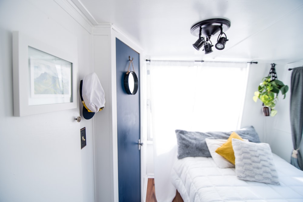 vier Kissen auf gestepptem weißem Bett in der Nähe der blauen Tür und des Fensters mit weißem Vorhang im gut beleuchteten Zimmer
