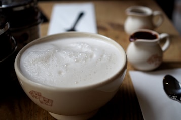 A frothy, creamy beverage fills a large bowl, with its smooth surface and bubbles clearly visible. In the background, two small creamers are placed on a wooden table, one of which appears to contain chocolate syrup. A spoon rests on a napkin nearby, and the soft lighting creates a warm, inviting atmosphere.