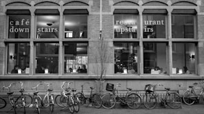 A black and white photo of a building's exterior with large windows displaying people sitting indoors. The windows have text indicating a café downstairs and a restaurant upstairs. Several bicycles are parked in front of the building.