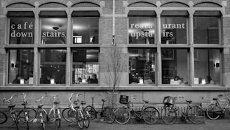 A black and white photo of a building's exterior with large windows displaying people sitting indoors. The windows have text indicating a café downstairs and a restaurant upstairs. Several bicycles are parked in front of the building.