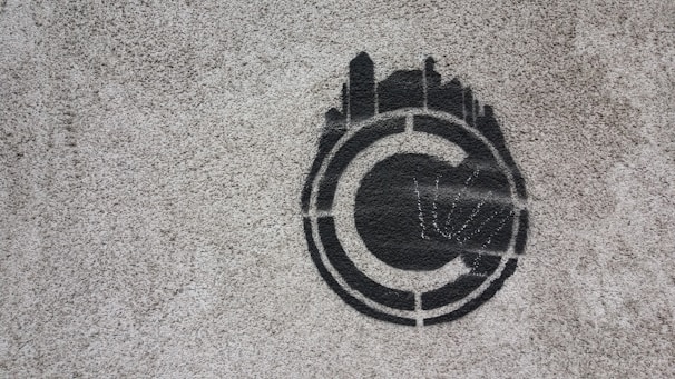 A stencil graffiti of a copyright symbol is painted on a rough concrete surface. The symbol is depicted in black with what appears to be skyline-like shapes emanating from the top half, giving it a distinctive and creative twist.