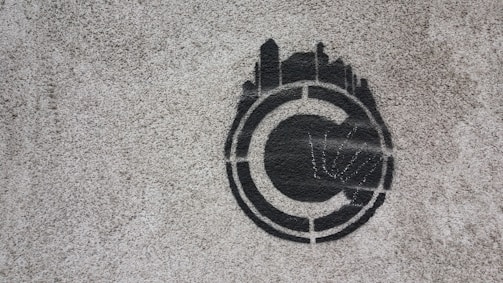 A stencil graffiti of a copyright symbol is painted on a rough concrete surface. The symbol is depicted in black with what appears to be skyline-like shapes emanating from the top half, giving it a distinctive and creative twist.