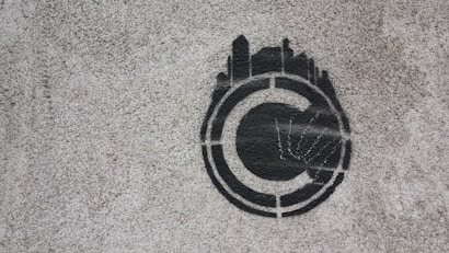 A stencil graffiti of a copyright symbol is painted on a rough concrete surface. The symbol is depicted in black with what appears to be skyline-like shapes emanating from the top half, giving it a distinctive and creative twist.