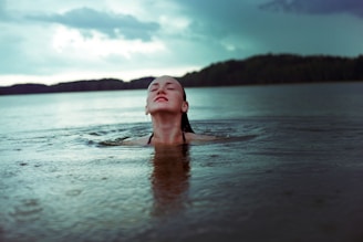 A person is emerging from the water in a calm lake, facing upwards with eyes closed. The background shows a serene landscape with a forested shoreline and a partly cloudy sky. The atmosphere is tranquil and reflective, suggesting a connection with nature.