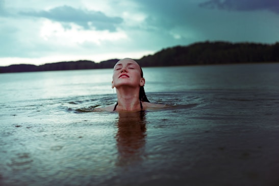 A person is emerging from the water in a calm lake, facing upwards with eyes closed. The background shows a serene landscape with a forested shoreline and a partly cloudy sky. The atmosphere is tranquil and reflective, suggesting a connection with nature.