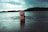 A person is emerging from the water in a calm lake, facing upwards with eyes closed. The background shows a serene landscape with a forested shoreline and a partly cloudy sky. The atmosphere is tranquil and reflective, suggesting a connection with nature.