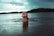 A person is emerging from the water in a calm lake, facing upwards with eyes closed. The background shows a serene landscape with a forested shoreline and a partly cloudy sky. The atmosphere is tranquil and reflective, suggesting a connection with nature.