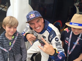 A man in a racing suit is posing enthusiastically with two children. The man is making a rock-and-roll hand gesture and wearing a cap with a Red Bull logo. One child is wearing a striped hoodie and a lanyard, while the other is in a T-shirt and straw hat with a similar lanyard.