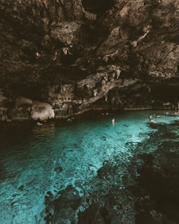 A serene cave pool with clear turquoise water reflecting the rugged rock ceiling overhead. Several people are swimming, enjoying the tranquility of the natural setting. Stalactites hang from the ceiling, adding a dramatic touch to the cave's ancient geology.