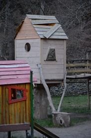 A wooden treehouse is built on a rustic structure with natural logs serving as supports. The house has a partially constructed roof and features a circular window and an open rectangular space. In the foreground is another smaller wooden structure with a red and yellow color scheme, featuring a triangular window.