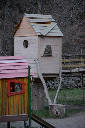A wooden treehouse is built on a rustic structure with natural logs serving as supports. The house has a partially constructed roof and features a circular window and an open rectangular space. In the foreground is another smaller wooden structure with a red and yellow color scheme, featuring a triangular window.