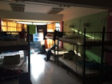 A dimly lit dormitory room with multiple bunk beds arranged along the walls. Light filters in through colorful curtains over a large set of windows, casting shadows on the tiled floor. The beds are made up with pillows and some personal belongings are scattered around, including backpacks on the floor and clothing on the beds.