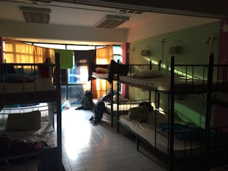 A dimly lit dormitory room with multiple bunk beds arranged along the walls. Light filters in through colorful curtains over a large set of windows, casting shadows on the tiled floor. The beds are made up with pillows and some personal belongings are scattered around, including backpacks on the floor and clothing on the beds.