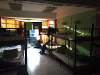 A dimly lit dormitory room with multiple bunk beds arranged along the walls. Light filters in through colorful curtains over a large set of windows, casting shadows on the tiled floor. The beds are made up with pillows and some personal belongings are scattered around, including backpacks on the floor and clothing on the beds.