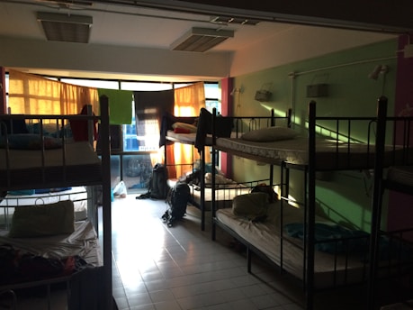 A dimly lit dormitory room with multiple bunk beds arranged along the walls. Light filters in through colorful curtains over a large set of windows, casting shadows on the tiled floor. The beds are made up with pillows and some personal belongings are scattered around, including backpacks on the floor and clothing on the beds.