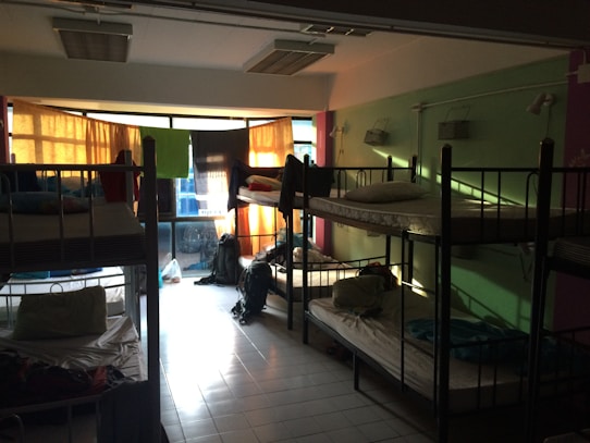 A dimly lit dormitory room with multiple bunk beds arranged along the walls. Light filters in through colorful curtains over a large set of windows, casting shadows on the tiled floor. The beds are made up with pillows and some personal belongings are scattered around, including backpacks on the floor and clothing on the beds.