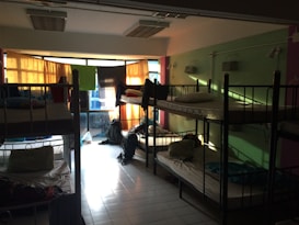 A dimly lit dormitory room with multiple bunk beds arranged along the walls. Light filters in through colorful curtains over a large set of windows, casting shadows on the tiled floor. The beds are made up with pillows and some personal belongings are scattered around, including backpacks on the floor and clothing on the beds.