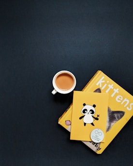 A warm cup of tea beside a stack of well-loved books and a curious cat peeking over.