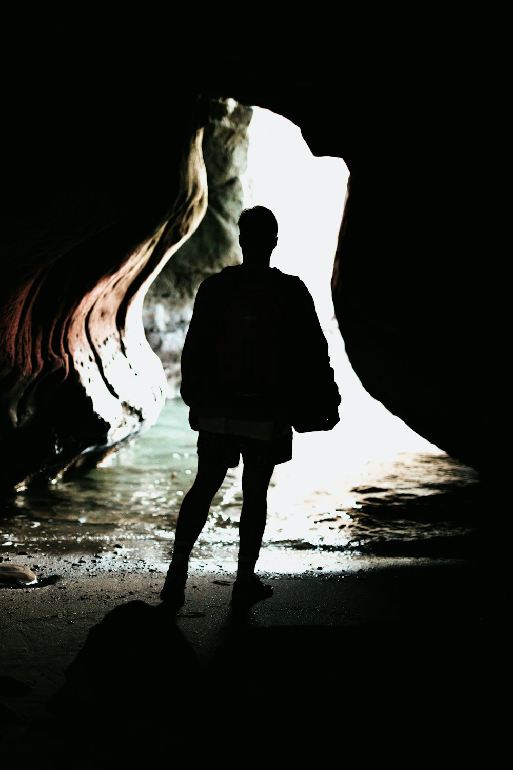 silhouette photo of person standing in front of blue body of water inside cave during daytime
