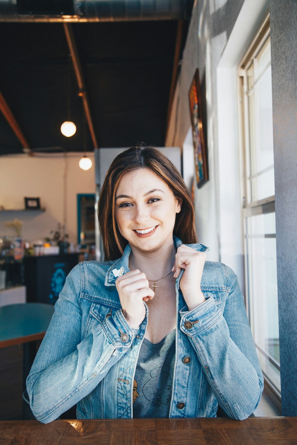 smiling woman wearing blue denim jacket holding her collar leaning on table beside clear glass window