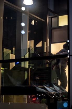 A dimly lit interior with a staircase and reflective surfaces. The image shows a person partially visible through a glass panel, surrounded by modern and abstract architectural elements. There are illuminated panels and geometric shapes creating a dynamic and contemporary atmosphere.