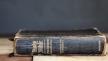 A worn, vintage edition of the Holy Bible is positioned horizontally on a wooden surface. The book's black cover displays signs of age, with frayed edges and visible wear. Gold embossed text can be seen on the spine and cover, indicating it is an analytical index edition by Dickson.