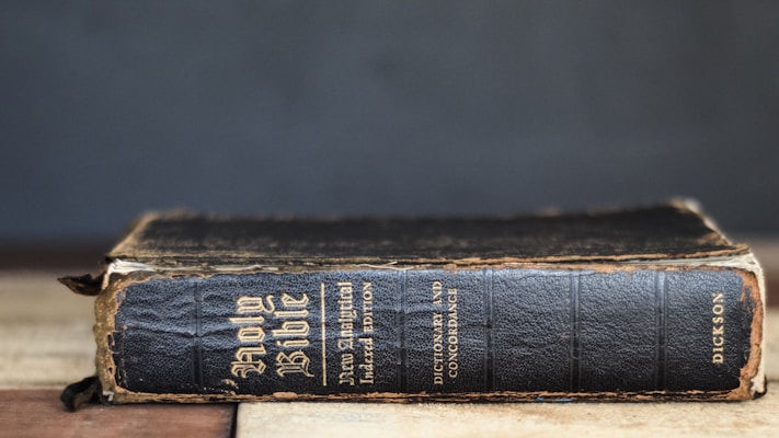 A worn, vintage edition of the Holy Bible is positioned horizontally on a wooden surface. The book's black cover displays signs of age, with frayed edges and visible wear. Gold embossed text can be seen on the spine and cover, indicating it is an analytical index edition by Dickson.